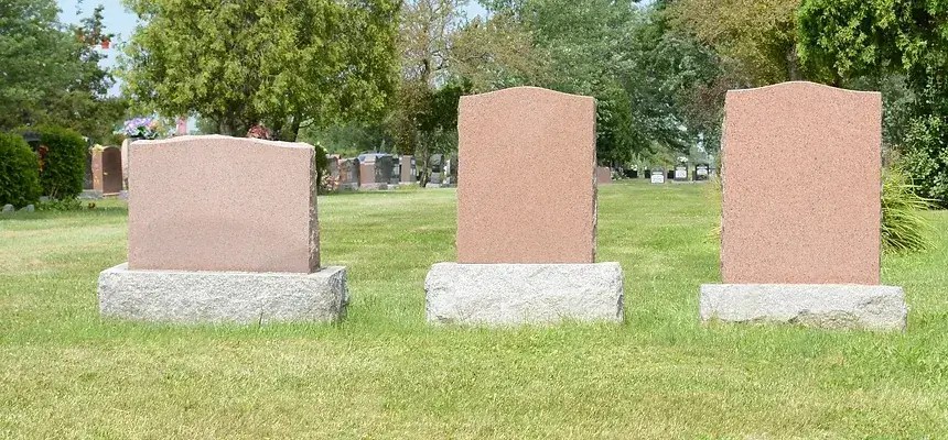 View of 3 different styles of headstones with headstones in the cemetery in the background out of focus.