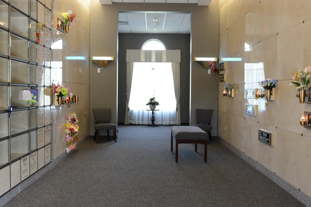 Inside view of cemetery mausoleum featuring a large window and crypts along the sides, covered in flowers.