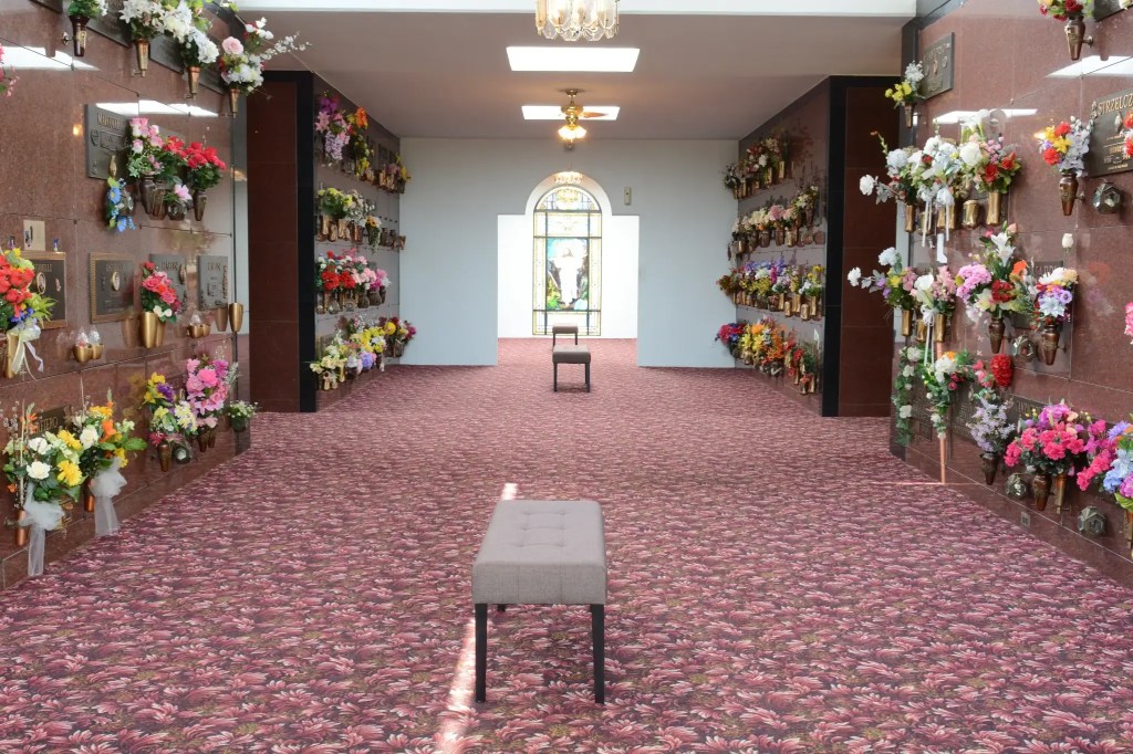 Inside view of cemetery mausoleum featuring a stained glass window and crypts along the sides, covered in flowers.