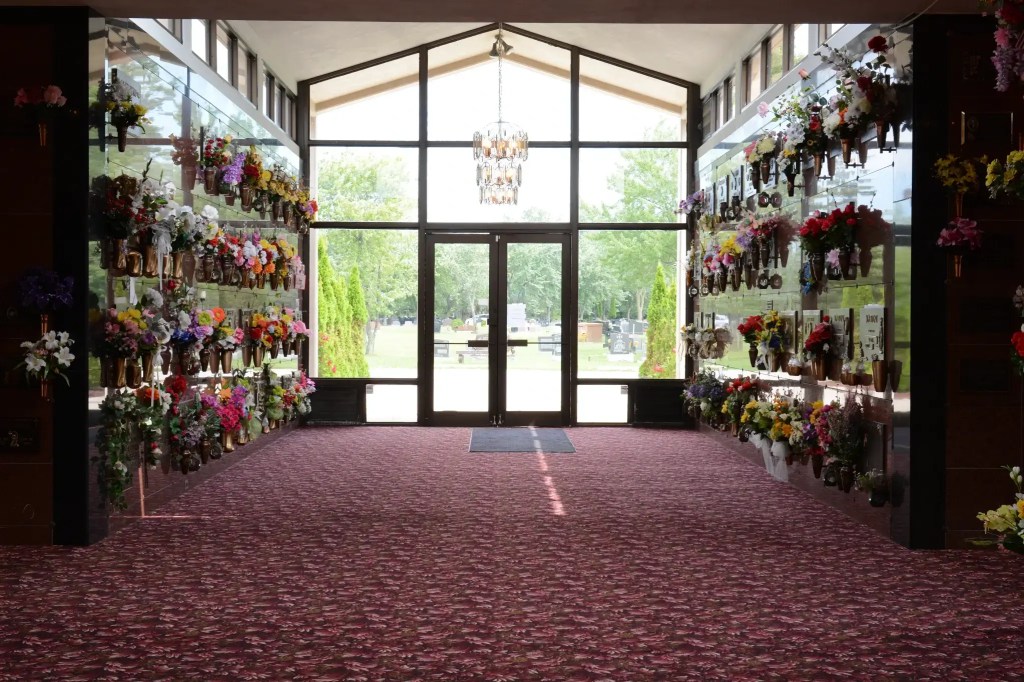 Inside view of cemetery mausoleum featuring a chandelier in front of a wall of windows. Crypts are along the sides, covered in flowers.