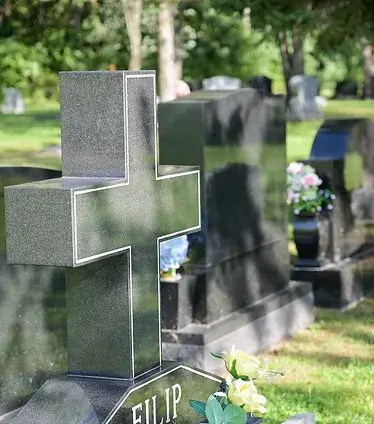 View of cross-shaped headstone with other headstones and floral arrangements in the cemetery out of focus in the background.