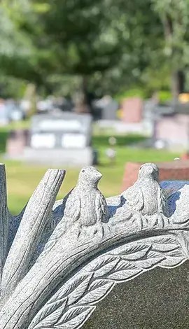 Closeup of headstone, featuring birds made of stone. Other headstones in the cemetery are visible in the background out of focus.